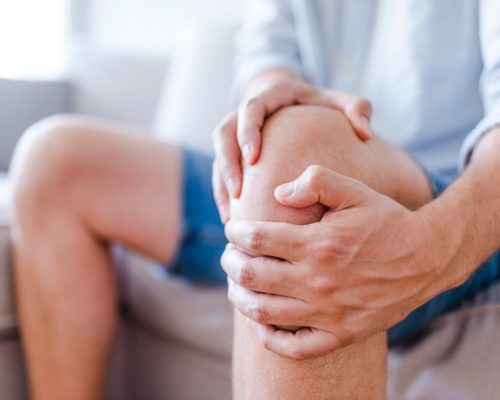 Man suffering from knee pain sitting sofa. A mature man massaging his painful knee. Man suffering from knee pain at home, closeup. Pain knee  (Man suffering from knee pain sitting sofa. A mature man massaging his painful knee. Man suffering from knee